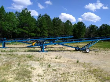 A blue metal conveyor system stands idle on a grassy, sandy surface, surrounded by trees under a clear, partly cloudy sky.
